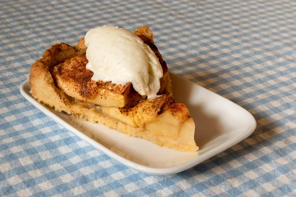 A wedge of apple pie with a scoop of vanilla ice cream on a gingham tablecloth.